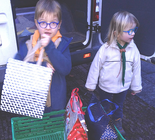 Ein Junge und ein Mädchen in Pfadfinderkleidung laden Geschenke aus einem Fahrzeug aus,