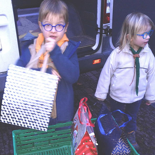 Ein Junge und ein Mädchen in Pfadfinderkleidung laden Geschenke aus einem Fahrzeug aus,