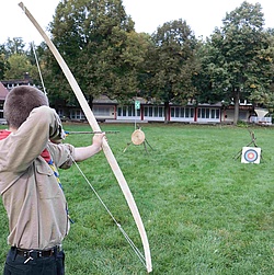 Ein Pfadfinder steht mit gespanntem Bogen vor einer Zielscheibe und zielt